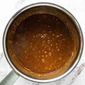 A saucepan filled with a thick brown sauce containing visible small bits, sitting on a white surface.