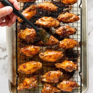 A hand uses a brush to glaze cooked chicken wings with sauce on a wire rack set over a baking sheet.