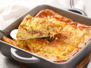 A square baking dish with a baked egg casserole; a slice is being lifted to show layers of egg, ham, and cheese.