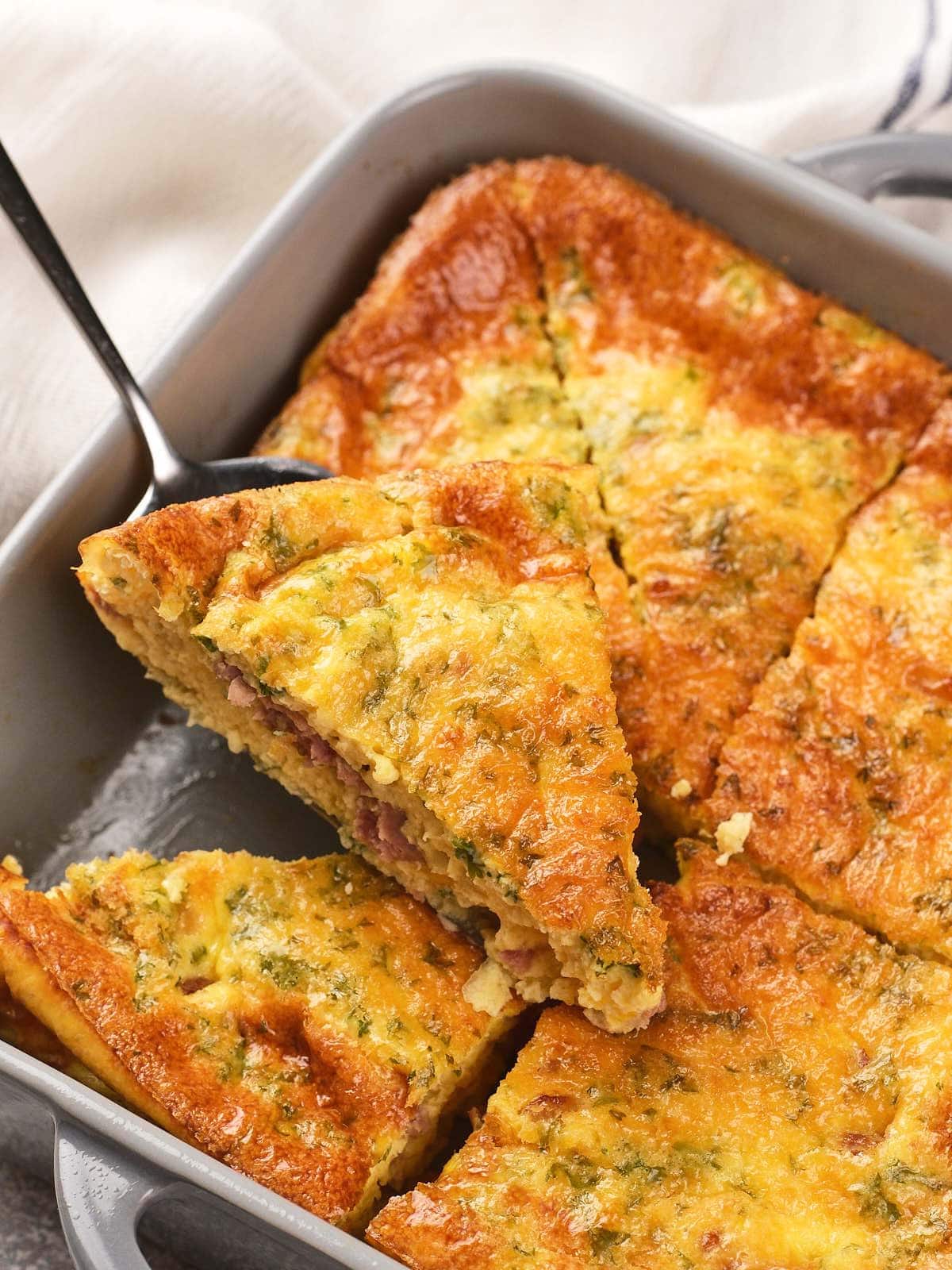 A close-up of a baked omelet with herbs and visible meat slices, cut into wedges in a square baking dish, with one wedge being lifted by a spatula.