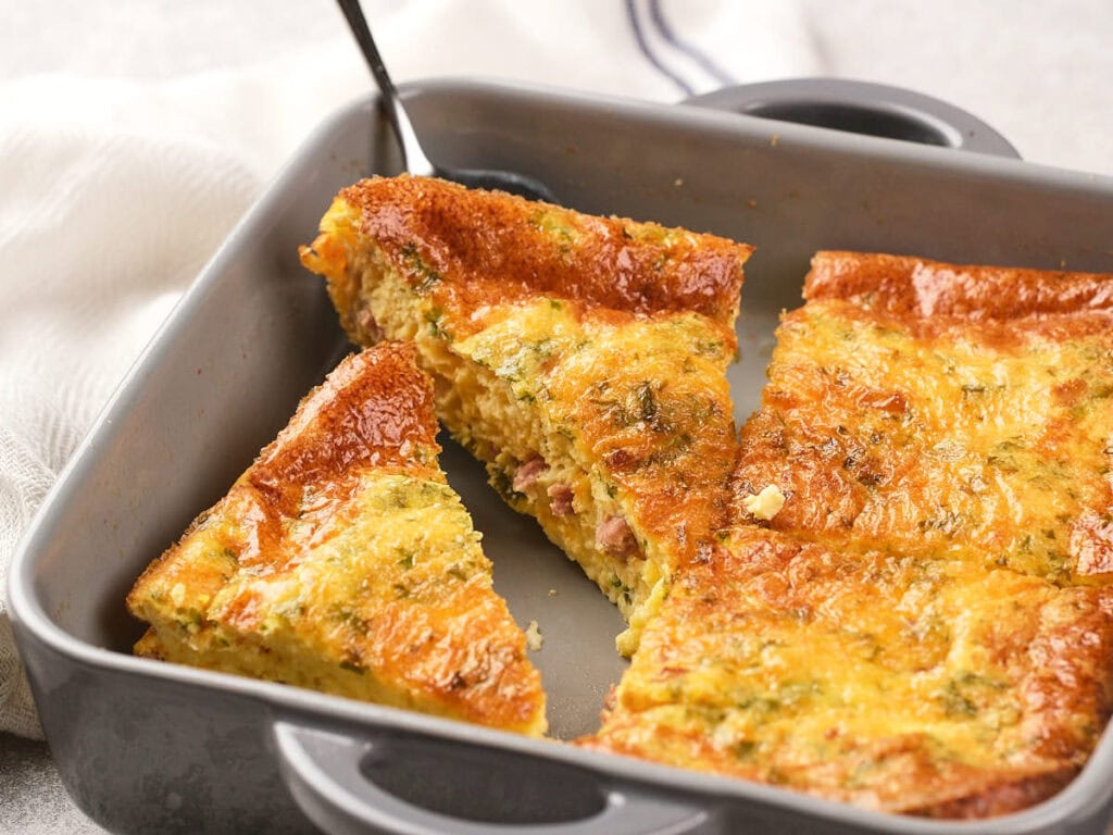 A baking pan with sliced baked omelet, featuring visible pieces of ham and herbs, with a serving spatula lifting one piece.