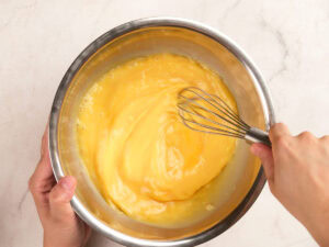 A person whisking a yellow mixture in a stainless steel bowl on a light-colored countertop.
