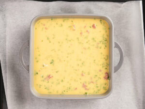 A square metal baking dish filled with an uncooked yellow egg mixture containing herbs and pieces of meat, placed on parchment paper.