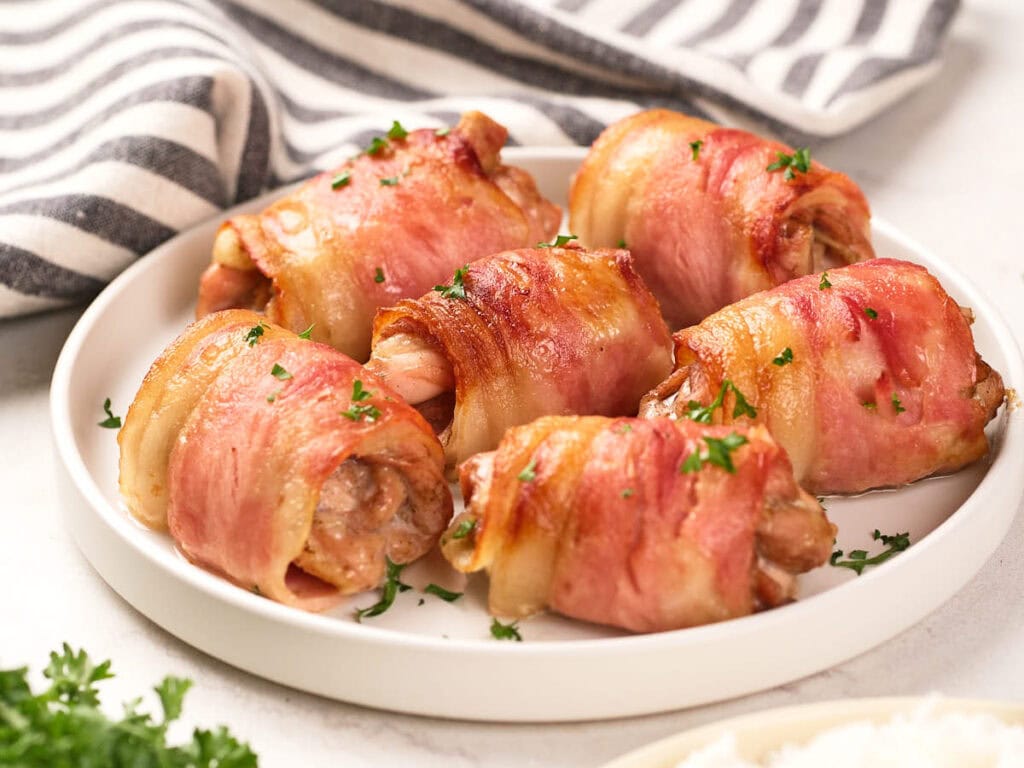 A white plate with six bacon-wrapped chicken rolls, garnished with chopped parsley. A striped cloth napkin is in the background.