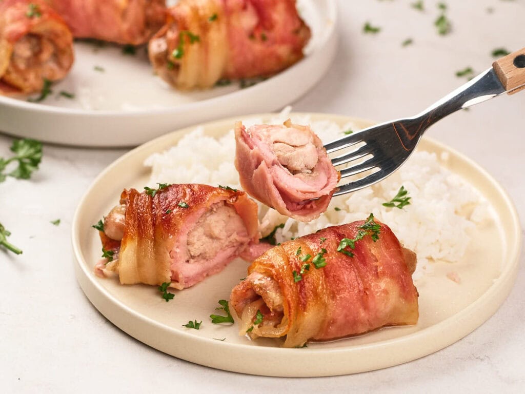 Bacon-wrapped chicken pieces garnished with parsley, served with white rice on a beige plate; one piece is held up on a fork.