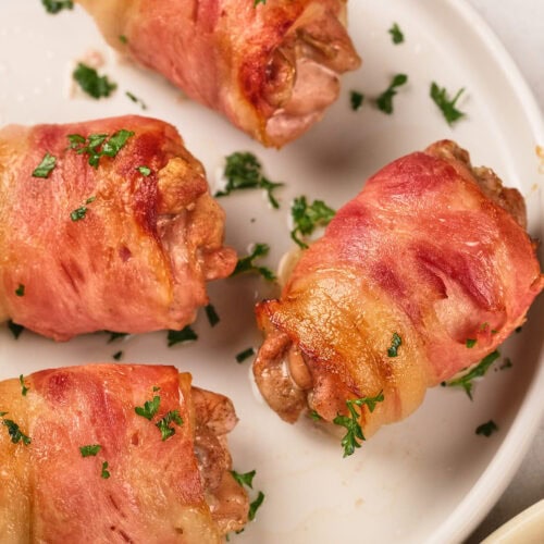 Four pieces of bacon-wrapped chicken garnished with chopped parsley are arranged on a white plate.