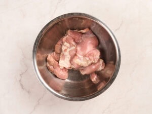 Raw pieces of chicken in a stainless steel mixing bowl on a light marble countertop.