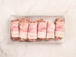Six pieces of bacon-wrapped meat are arranged in a row on a rectangular white dish, placed on a light marble surface.