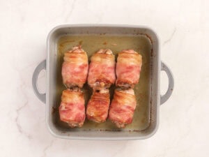 Six pieces of bacon-wrapped chicken are arranged in two rows inside a square baking dish on a light-colored surface.