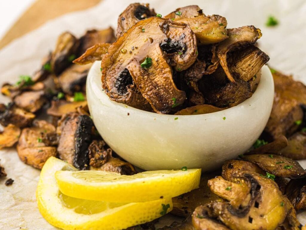 Sliced sautéed mushrooms garnished with parsley in a small white bowl, with lemon wedges on the side.