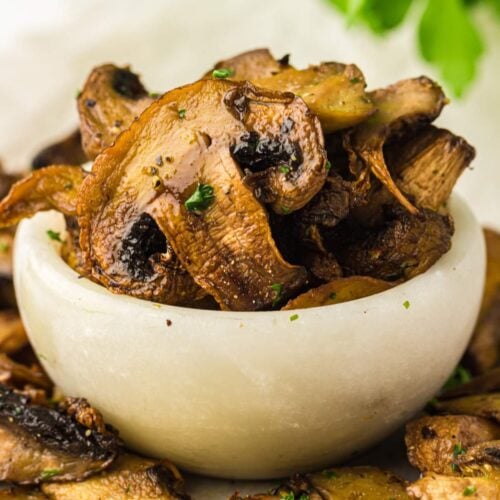 A small white bowl filled with cooked, sliced mushrooms, garnished with herbs, with fresh parsley and a lemon slice nearby.