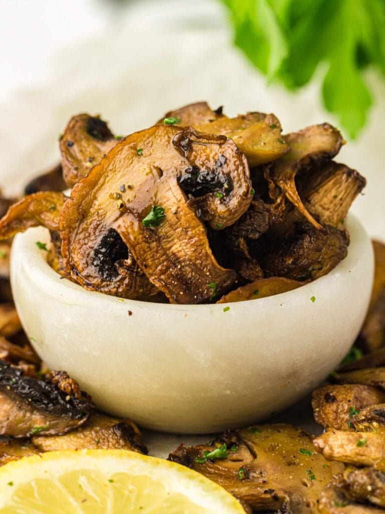 A small white bowl filled with cooked, sliced mushrooms, garnished with herbs, with fresh parsley and a lemon slice nearby.