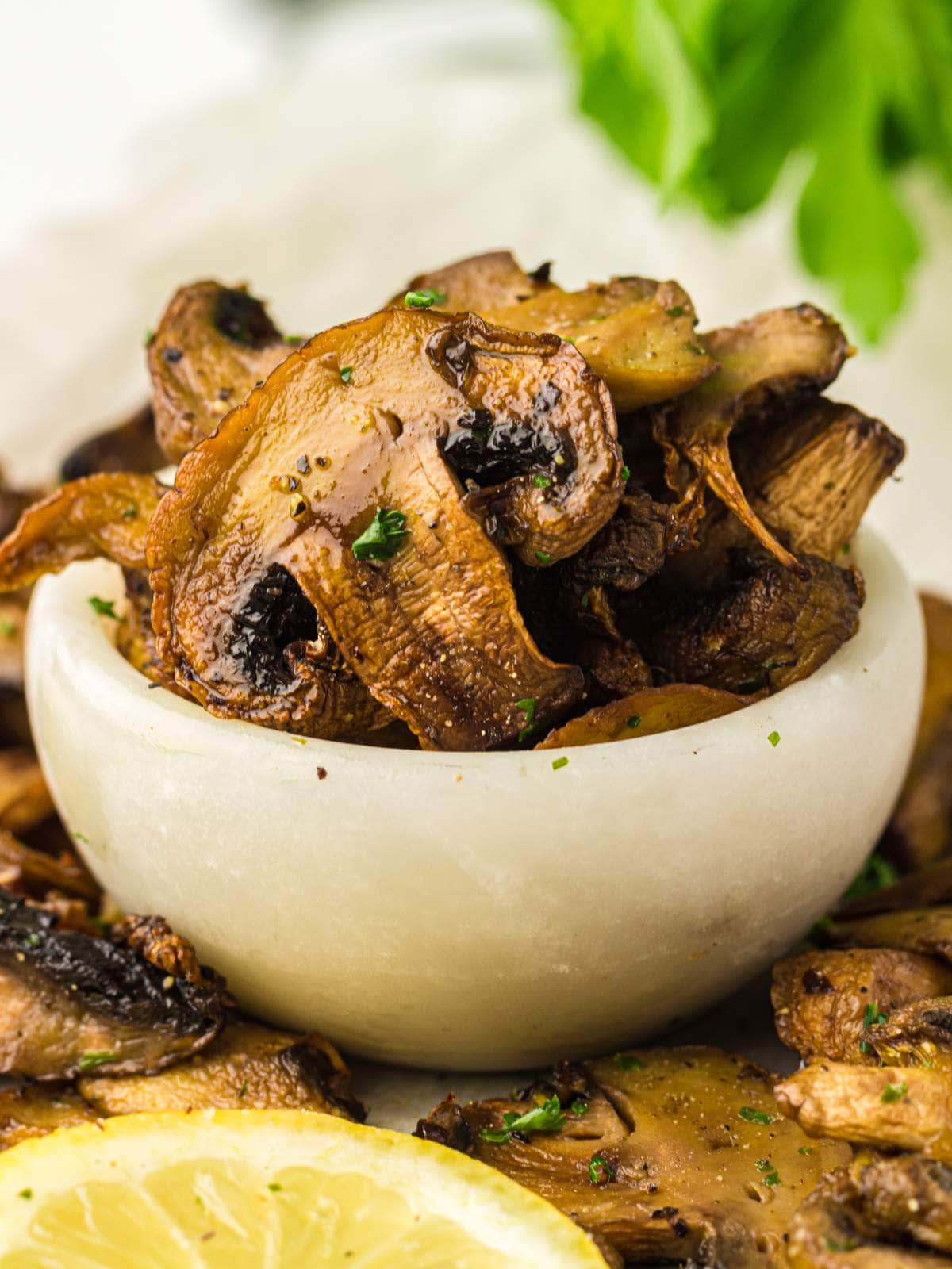 A small white bowl filled with cooked, sliced mushrooms, garnished with herbs, with fresh parsley and a lemon slice nearby.