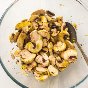 Sliced mushrooms mixed with herbs and oil in a clear glass bowl, with a metal spoon on the side.