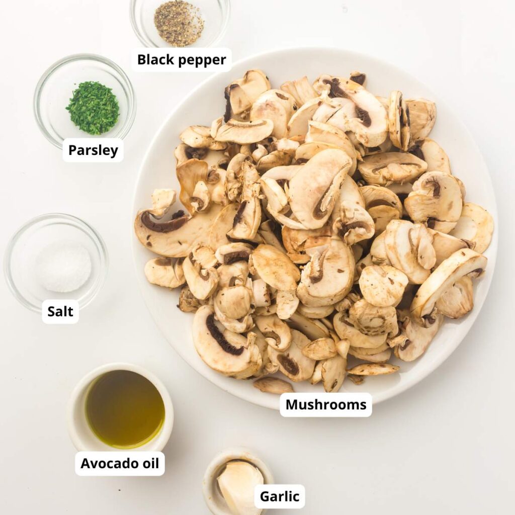 A plate of sliced mushrooms surrounded by bowls of parsley, black pepper, salt, avocado oil, and garlic, all labeled.
