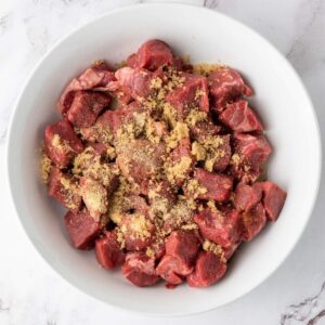 Chunks of raw beef in a white bowl, topped with a sprinkling of brown sugar and seasonings, on a white marble surface.