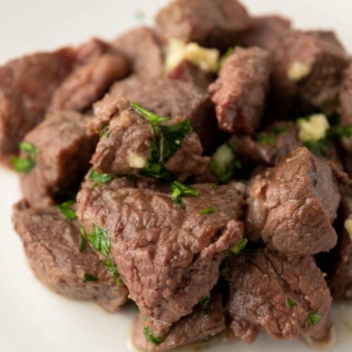 A plate of cooked beef cubes garnished with chopped herbs and small pieces of garlic on a white background.