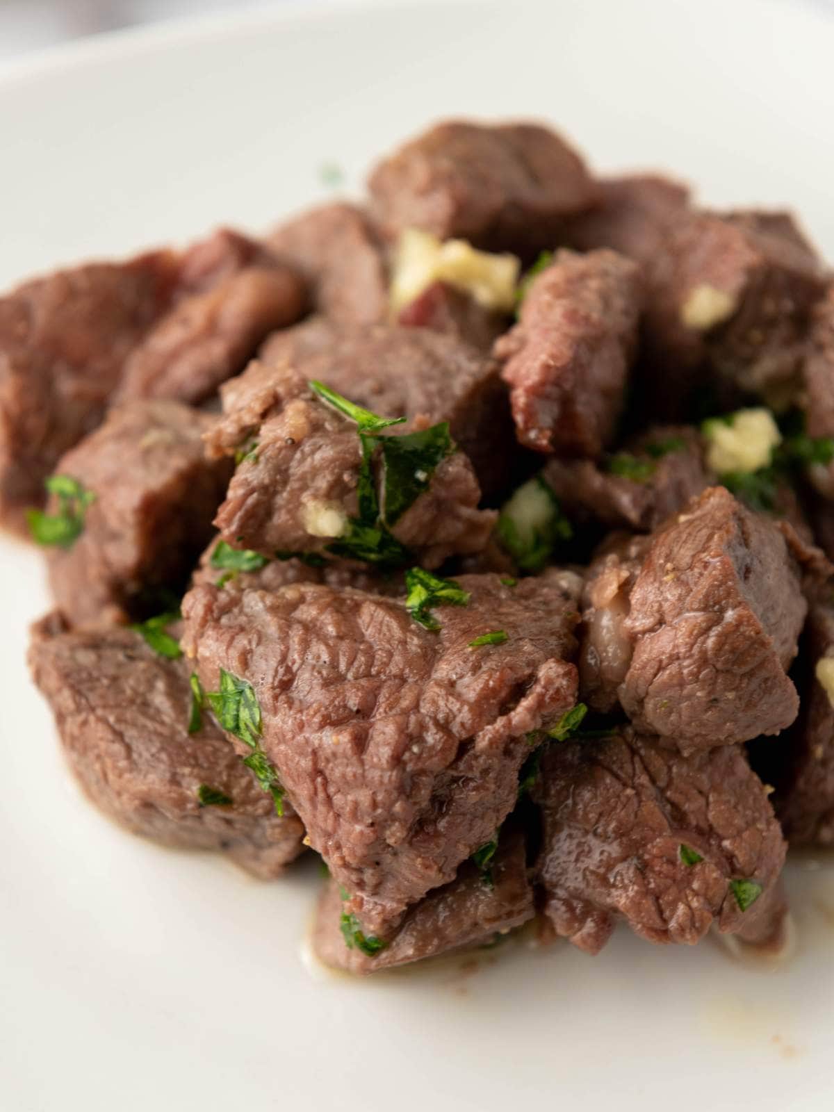 A plate of cooked beef cubes garnished with chopped herbs and small pieces of garlic on a white background.