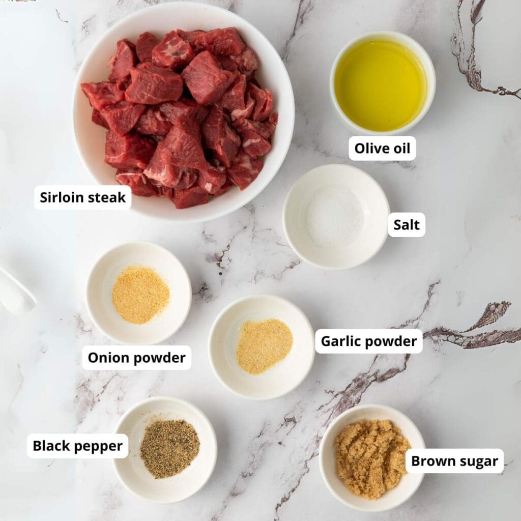 Bowls containing sirloin steak, olive oil, salt, onion powder, garlic powder, black pepper, and brown sugar are arranged on a white marble surface. Each ingredient is labeled.