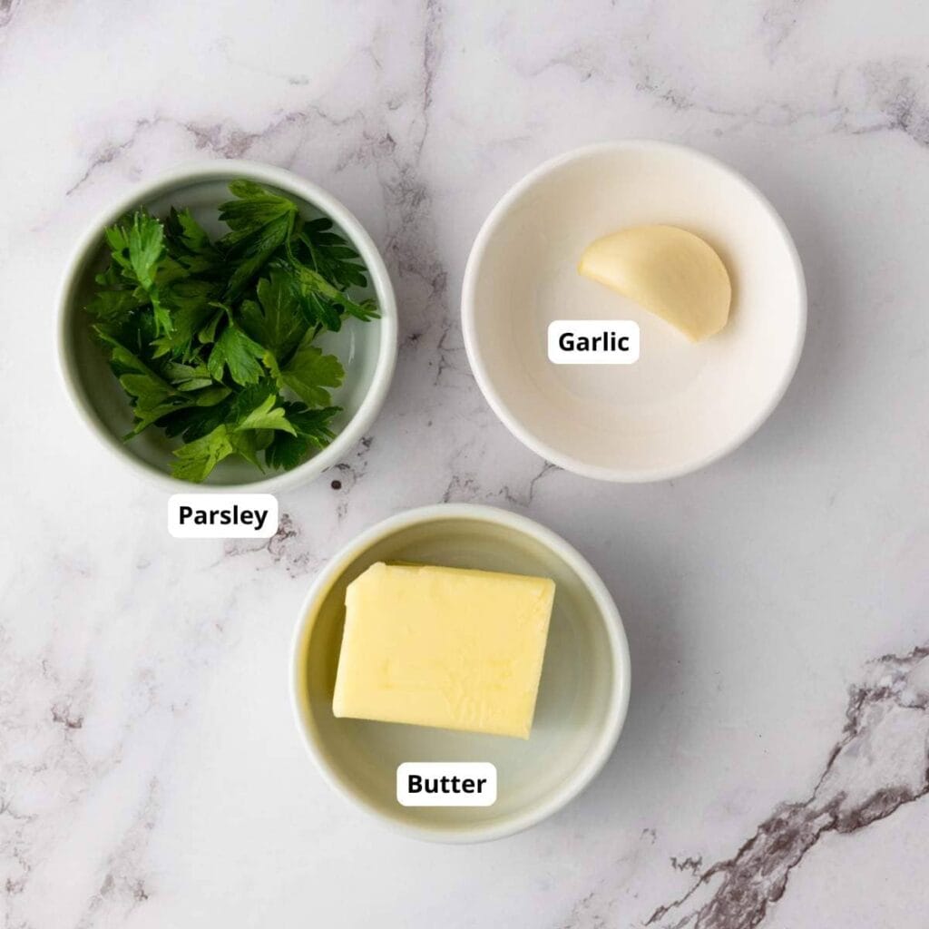 Three small bowls on a marble surface containing fresh parsley, a clove of garlic, and a piece of butter, each labeled.