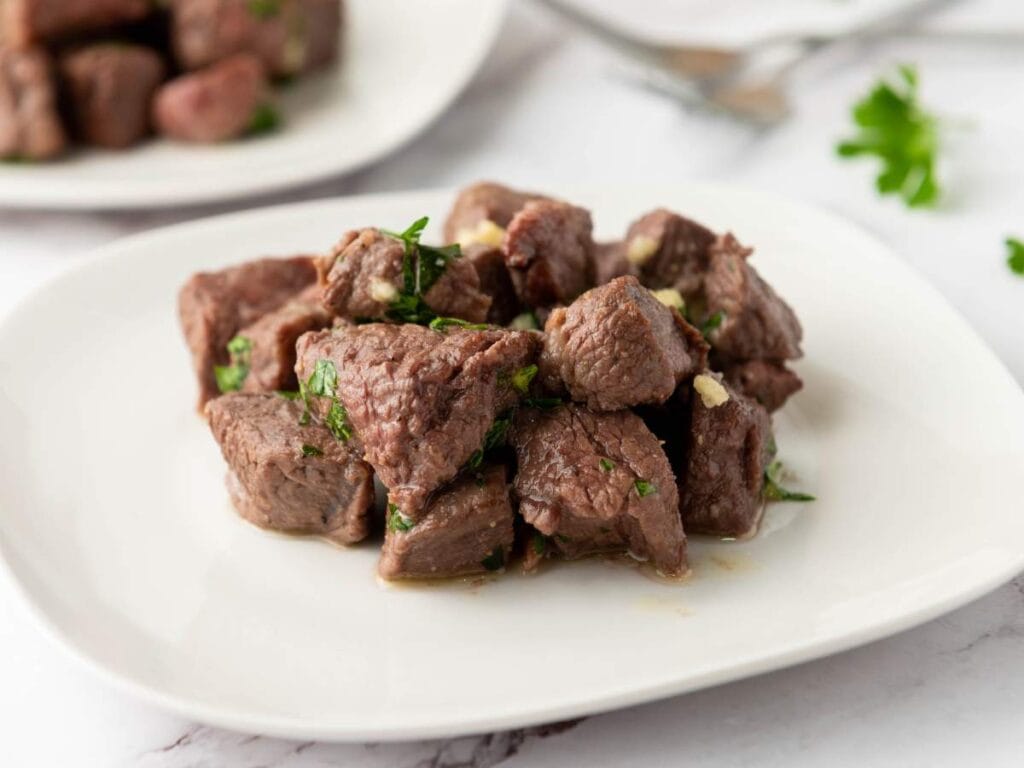 A white plate with cooked beef steak bites garnished with chopped herbs, placed on a light surface.