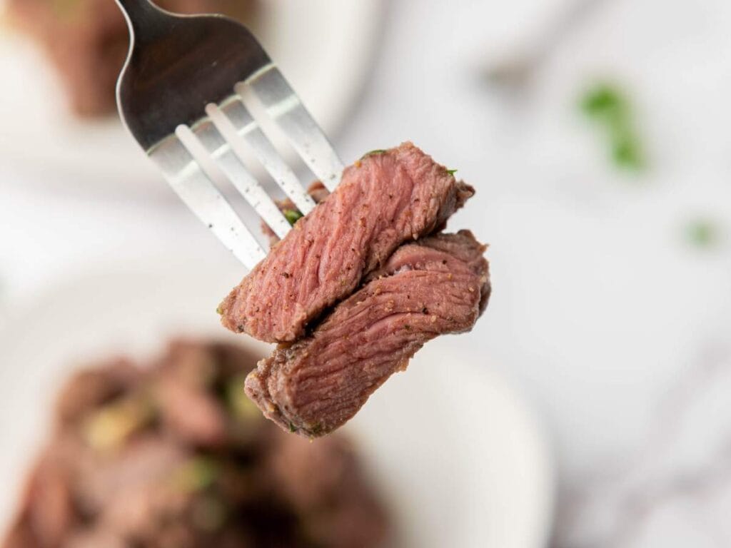 A close-up of two pieces of cooked steak on a fork, with a blurred plate of more steak in the background.