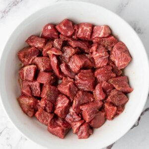 A white bowl filled with raw, cubed pieces of red meat on a marble surface.