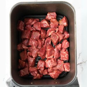Chunks of raw beef placed in a single layer inside an air fryer basket.
