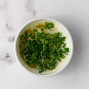 A white bowl filled with melted butter topped with freshly chopped green herbs, placed on a light marble surface.