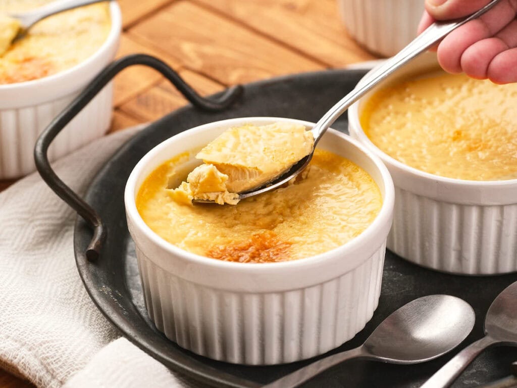 A hand holds a spoonful of creamy custard from a white ramekin on a tray, with another ramekin and spoons nearby.