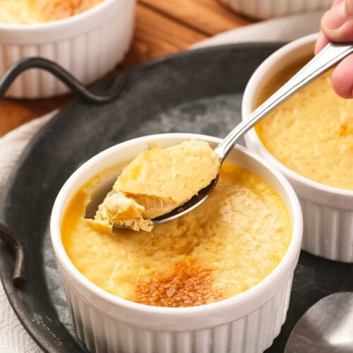A hand holding a spoon lifts a portion of baked custard from a white ramekin, with more ramekins and a spoon on a dark tray.