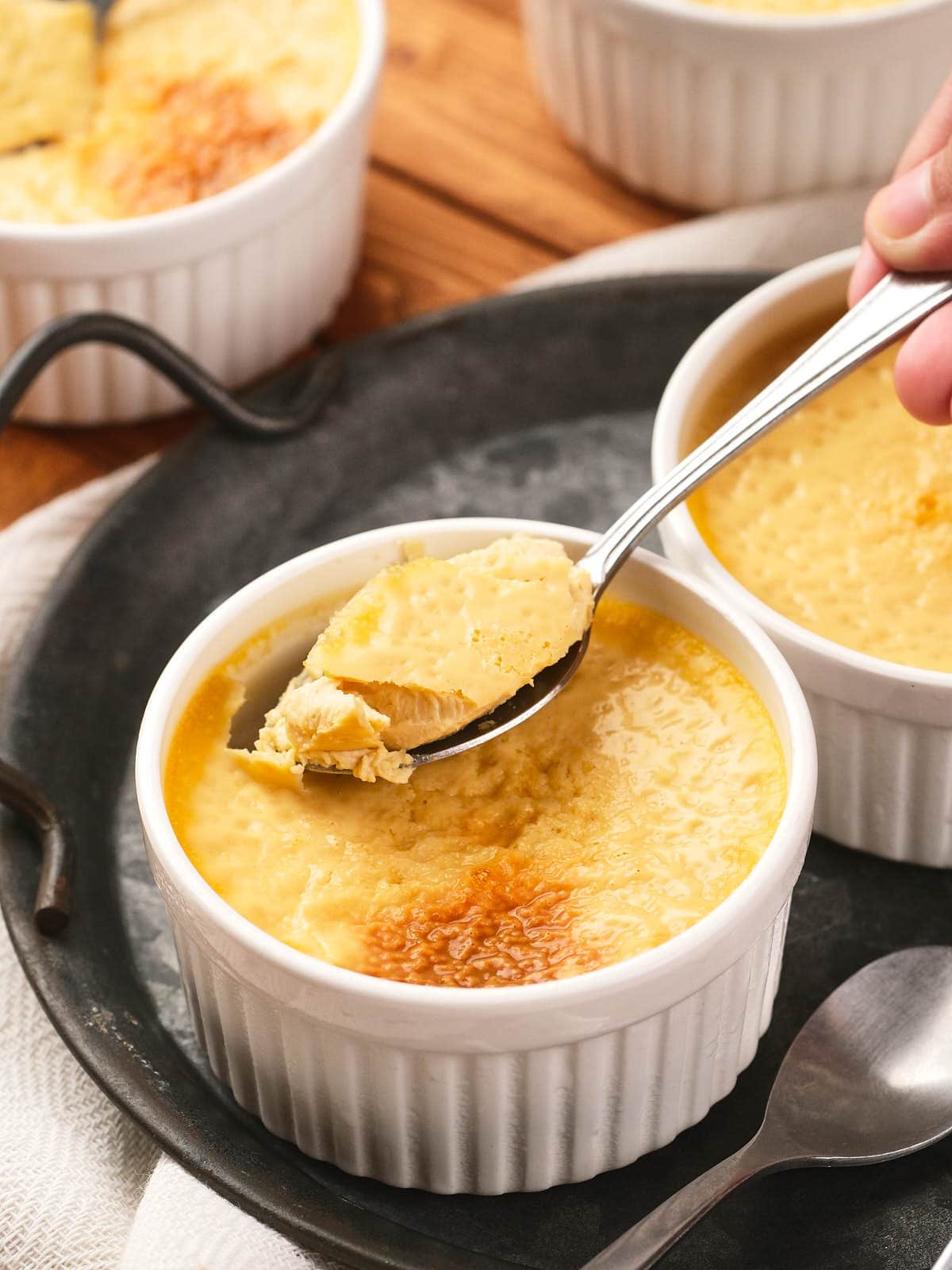 A hand holding a spoon lifts a portion of baked custard from a white ramekin, with more ramekins and a spoon on a dark tray.
