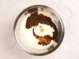 A stainless steel bowl containing cream, brown sugar, molasses, an egg, and vanilla extract on a light countertop.