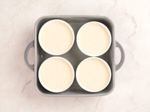Four white ramekins filled with cream-colored custard mixture are placed in a gray baking pan on a light marble surface.