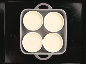Four white ramekins filled with a light-colored mixture are placed in a square baking dish on a black tray, viewed from above.