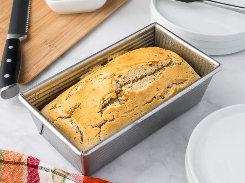 A loaf of bread with a cracked top sits in a metal loaf pan on a countertop, next to a knife, plates, and a colorful cloth.