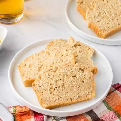 Three slices of light brown bread are arranged on a white plate, with another plate of bread, a drink, and a colorful checkered cloth nearby.