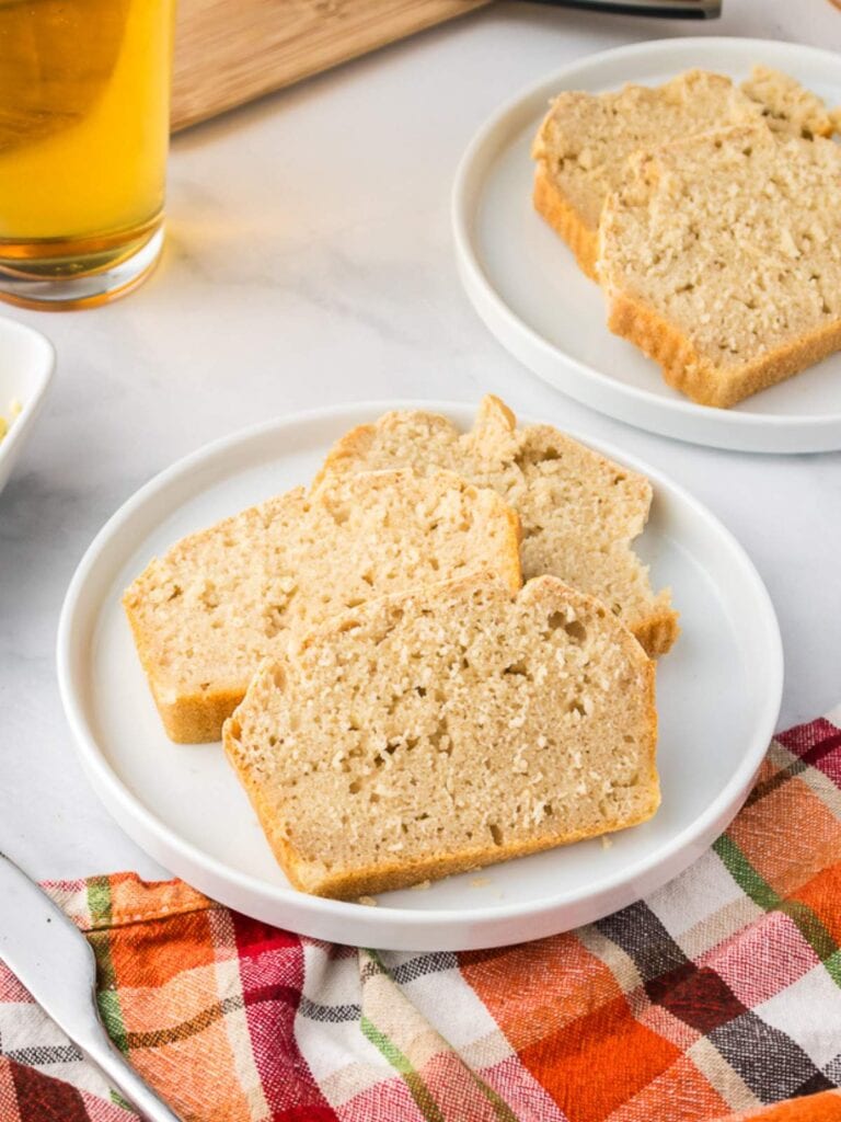 Three slices of light brown bread are arranged on a white plate, with another plate of bread, a drink, and a colorful checkered cloth nearby.