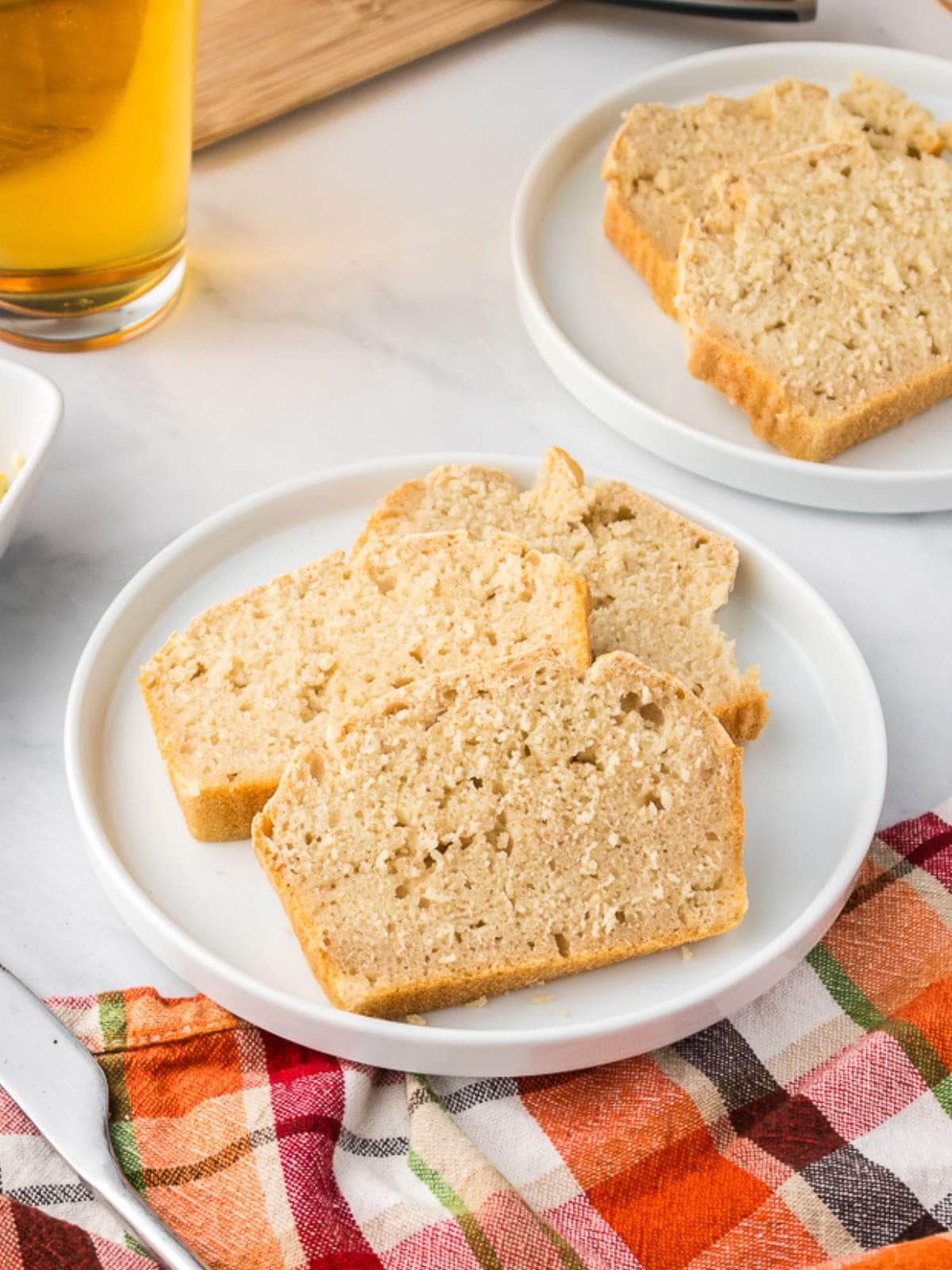 Three slices of light brown bread are arranged on a white plate, with another plate of bread, a drink, and a colorful checkered cloth nearby.
