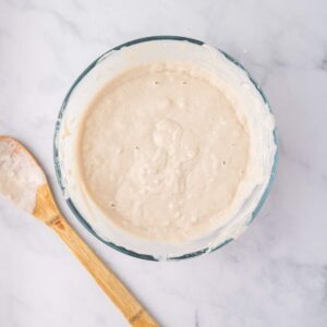 A glass bowl filled with thick, creamy batter sits on a marble surface next to a wooden spoon coated with batter.