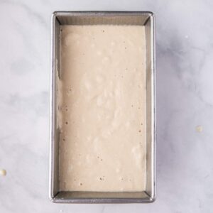 A metal loaf pan filled with pale, bubbly bread batter sits on a light marble surface.