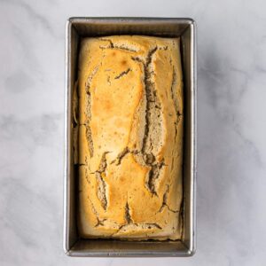 A loaf of bread with a cracked, golden-brown crust sits in a rectangular metal baking pan on a light, marbled surface.