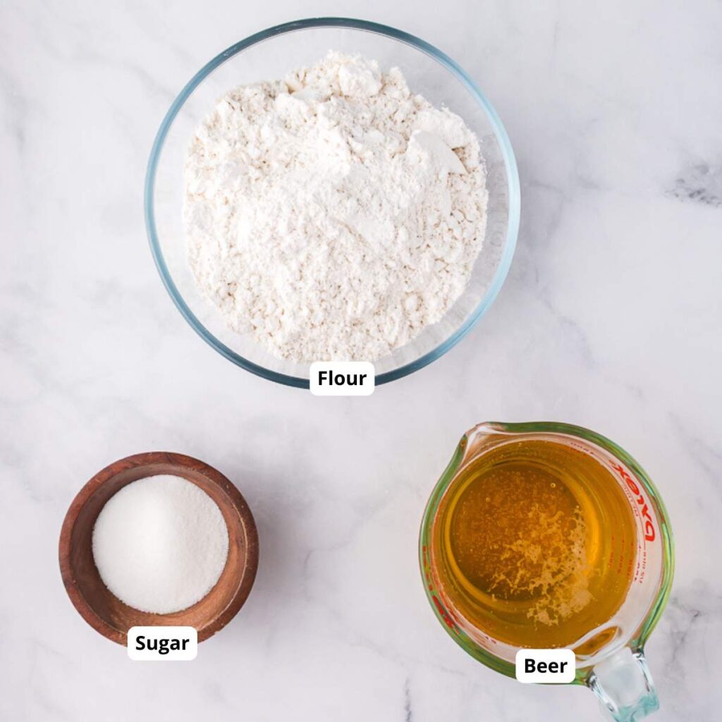 A glass bowl of flour, a wooden bowl of sugar, and a measuring cup of beer are arranged on a marble surface, each labeled accordingly.
