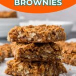 A stack of three coconut butterscotch brownies on parchment paper, with more brownies and a plate visible in the background.