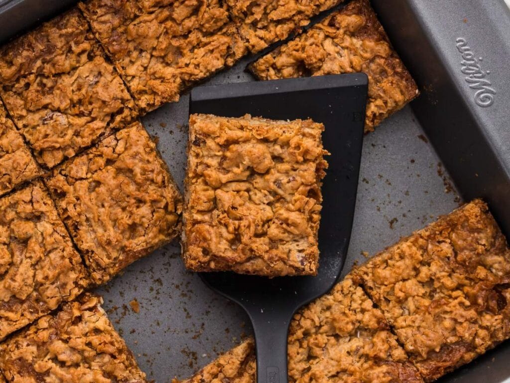 A baking pan of butterscotch brownies cut into squares, with one bar lifted out on a black spatula.