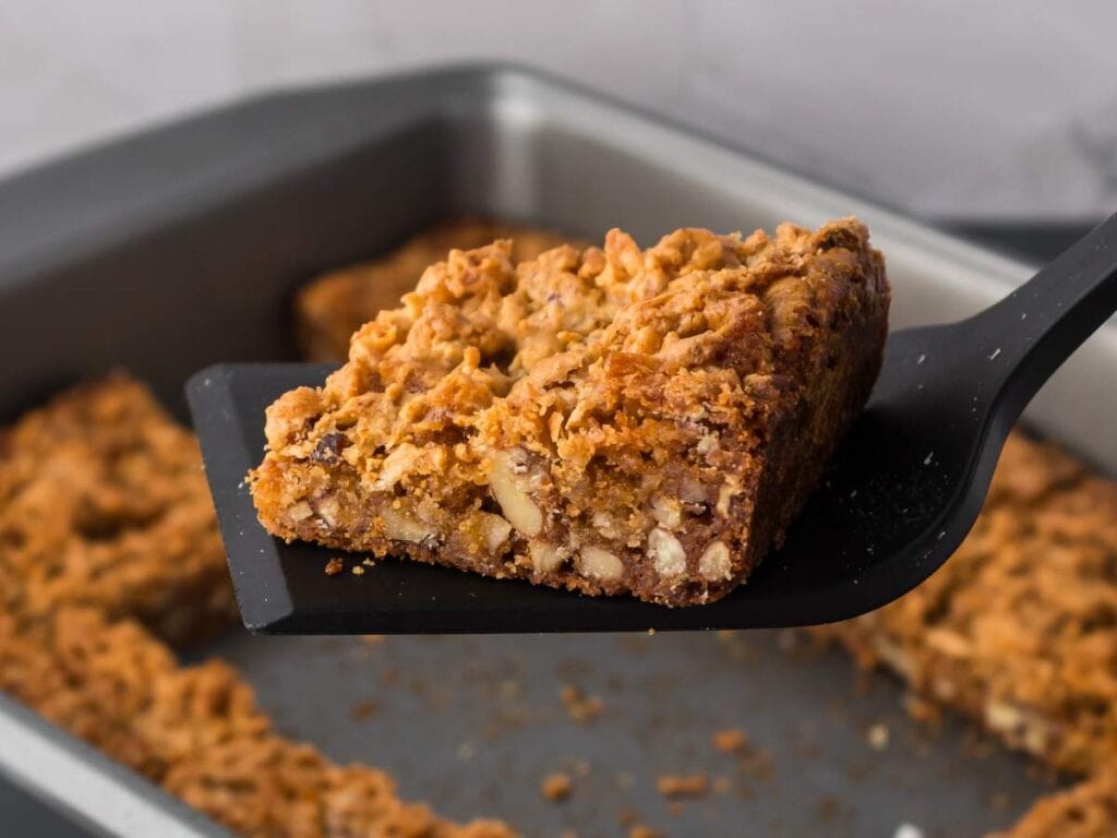 A close-up of a brownie bar with visible nuts on a black spatula, held above a baking pan containing more blondie bars.