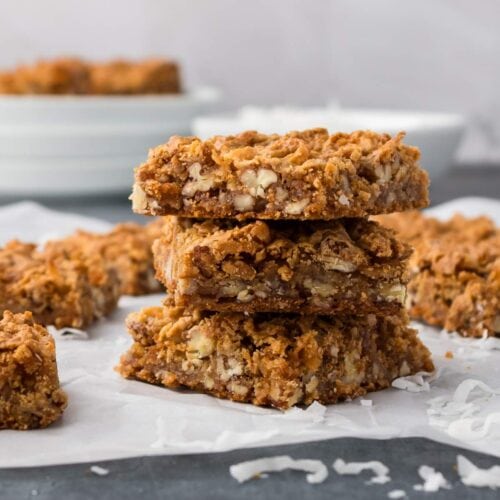 Three stacked oat and nut bars are placed on parchment paper, with additional bars and shredded coconut scattered around on a dark surface.