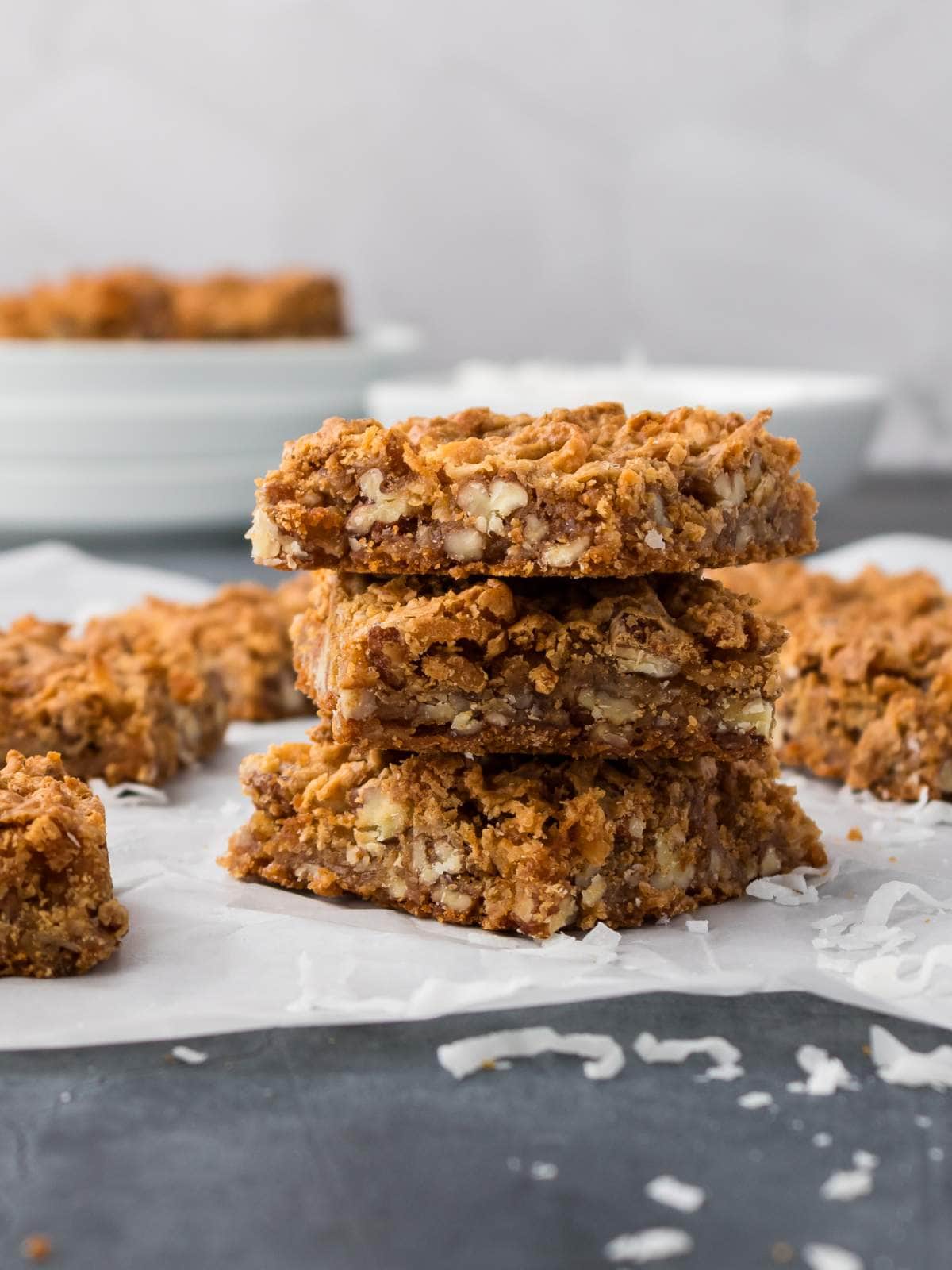 Three stacked oat and nut bars are placed on parchment paper, with additional bars and shredded coconut scattered around on a dark surface.