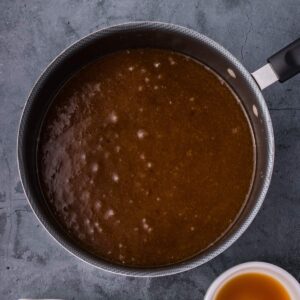 A saucepan filled with a thick brown liquid, possibly a sauce or gravy, on a dark countertop.