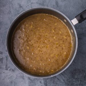 A saucepan filled with a brown, simmering liquid mixture, viewed from above, on a gray textured surface.